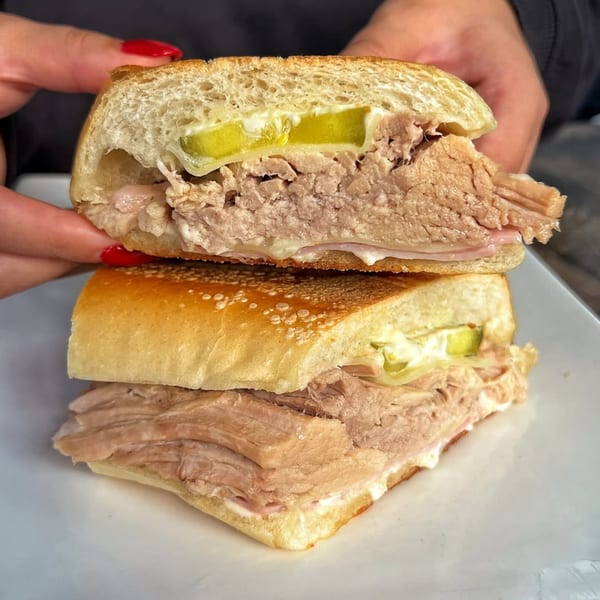 Hands holding a sliced Dominican Cuban sandwich with pork, ham, cheese, pickles, and mayo.
