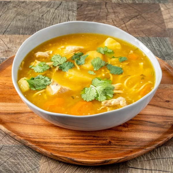 Dominican chicken soup with noodles, potatoes, carrots, and cilantro in a white bowl on a wooden tray.