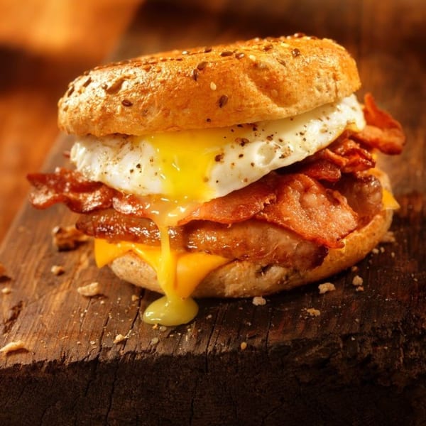A breakfast bagel sandwich with a runny fried egg, crispy bacon, and melted cheese on a wooden board.