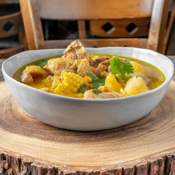 Steaming Dominican sancocho, a hearty stew with meat, corn, and potatoes, in a white bowl.
