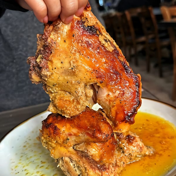 A hand holds up a piece of golden-brown roasted chicken over a plate with more chicken and sauce.